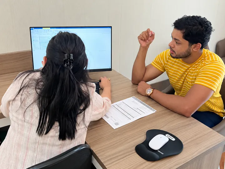 Suzane Salvador ao computador, de costas, atendendo um cliente sentado à sua frente, com uma guia sobre a mesa